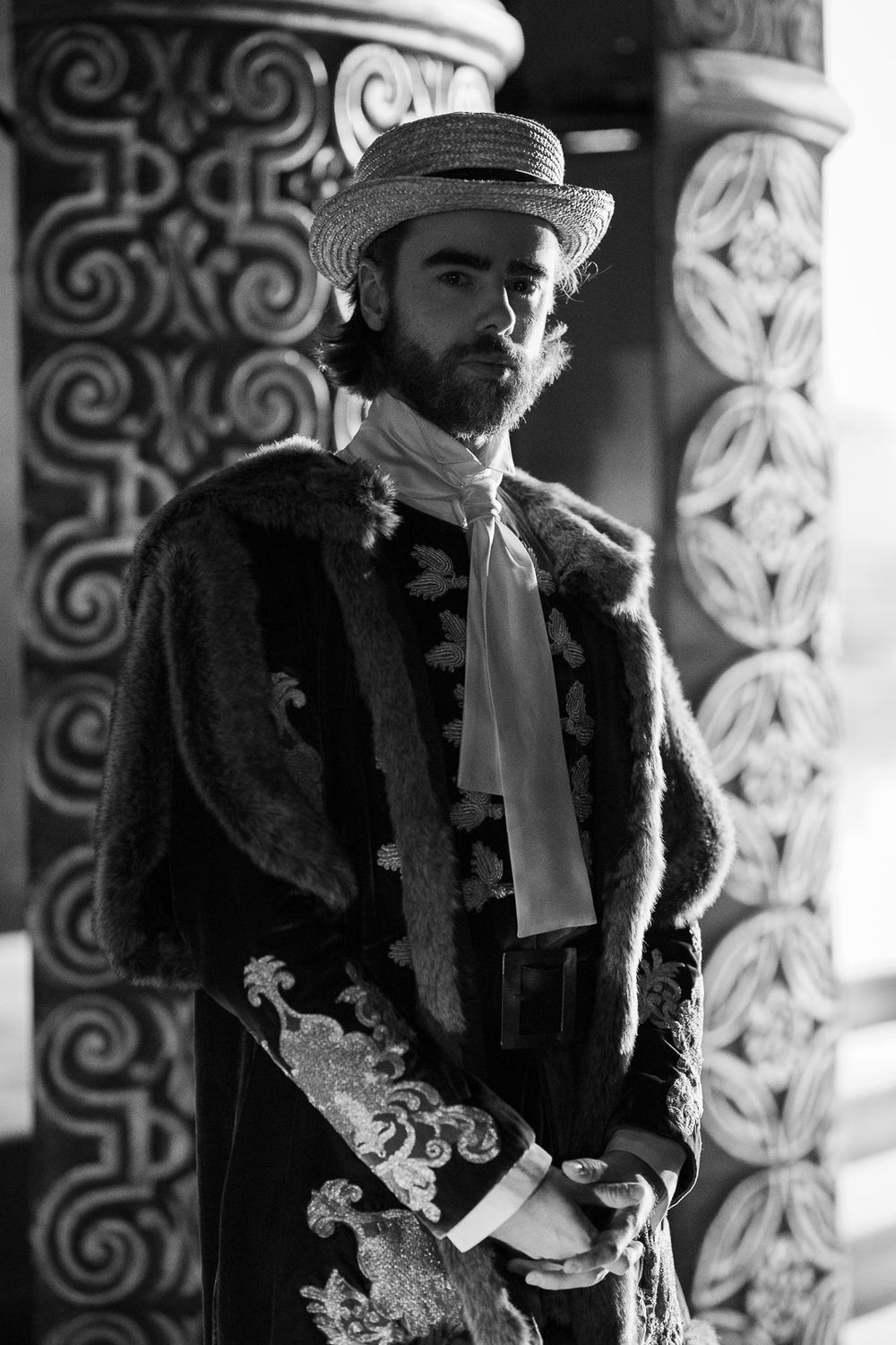 Black and white portrait of a bearded man in ornate historical costume with straw hat beside decorative columns.