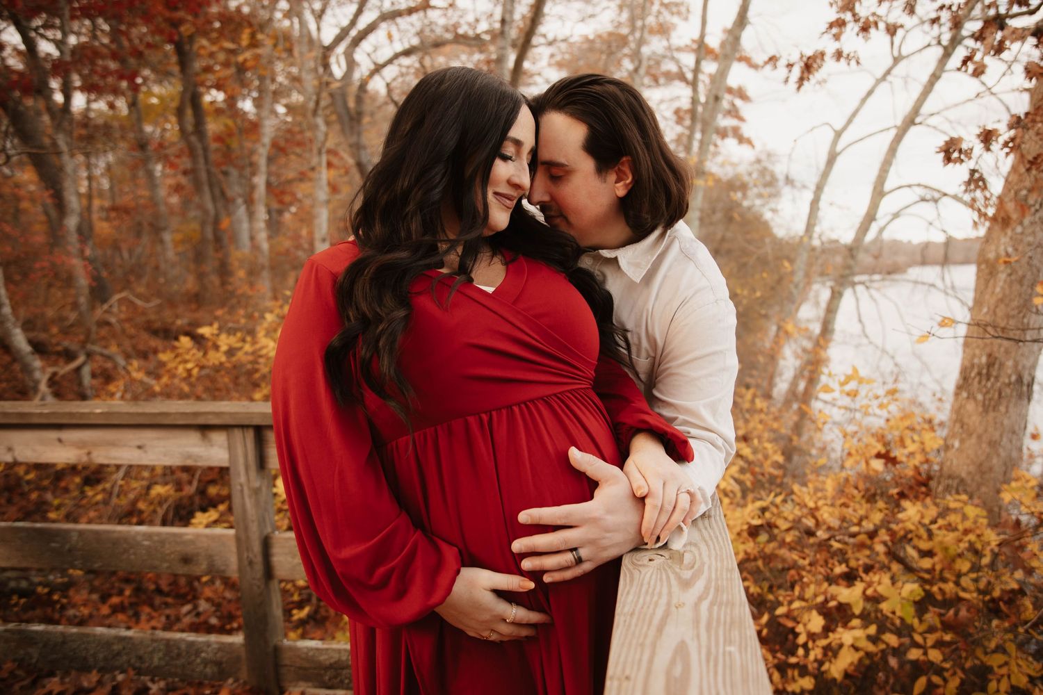 Couple embracing outdoors during fall maternity photoshoot, woman in red dress surrounded by autumn foliage.