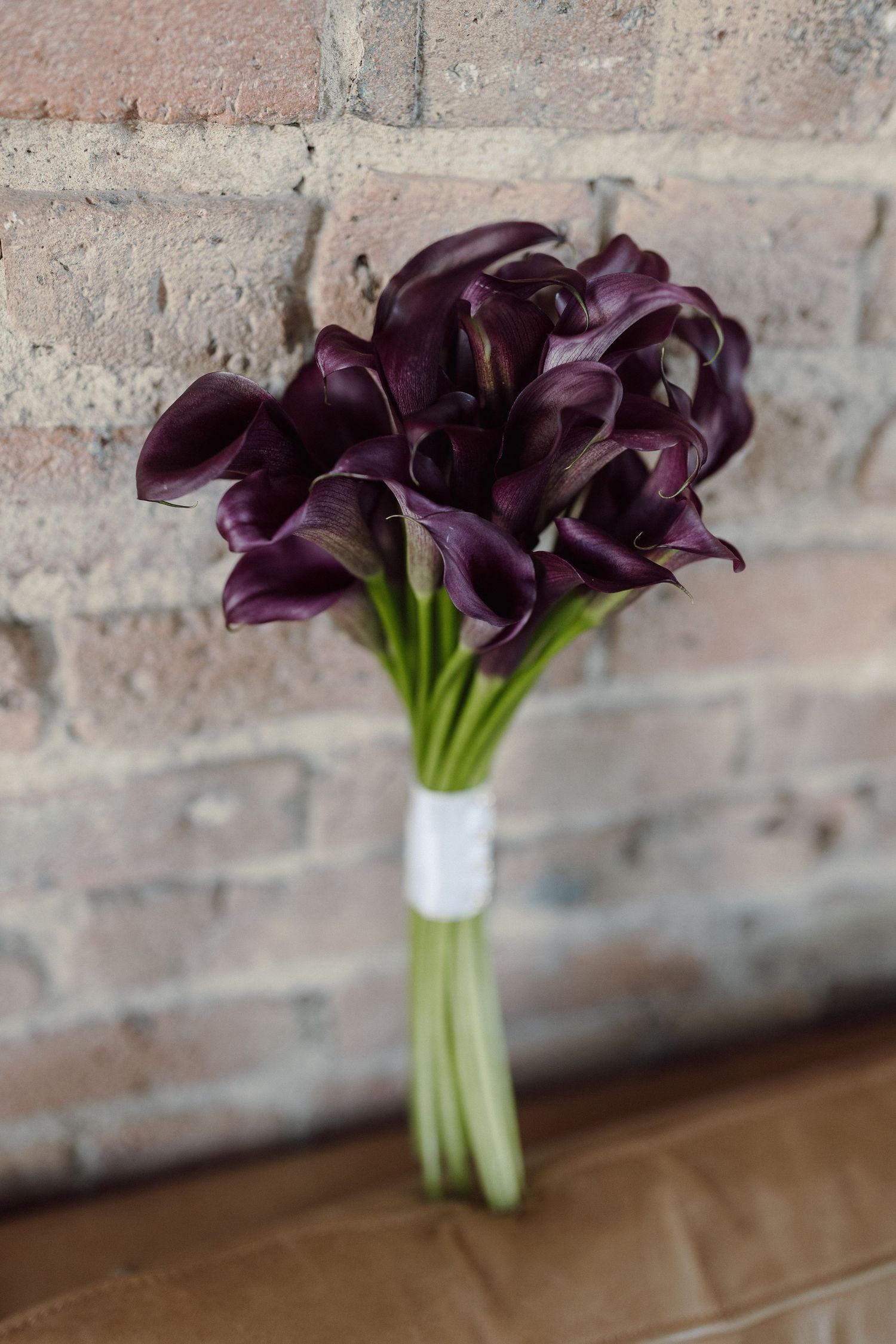 Deep purple calla lily bouquet arranged against a rustic brick wall.