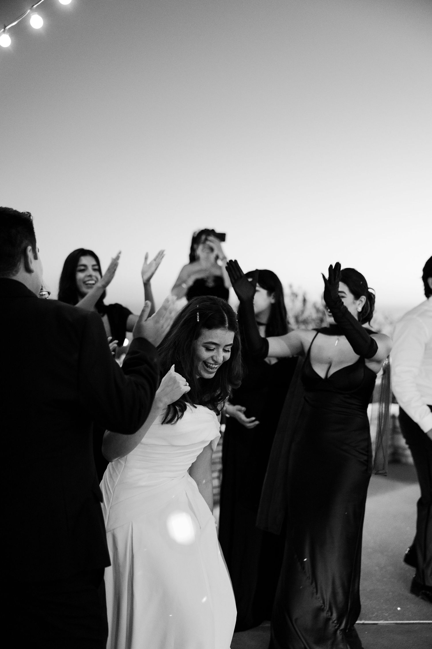 A black and white photo of wedding guests dancing and celebrating at a reception with the bride in a white dress.