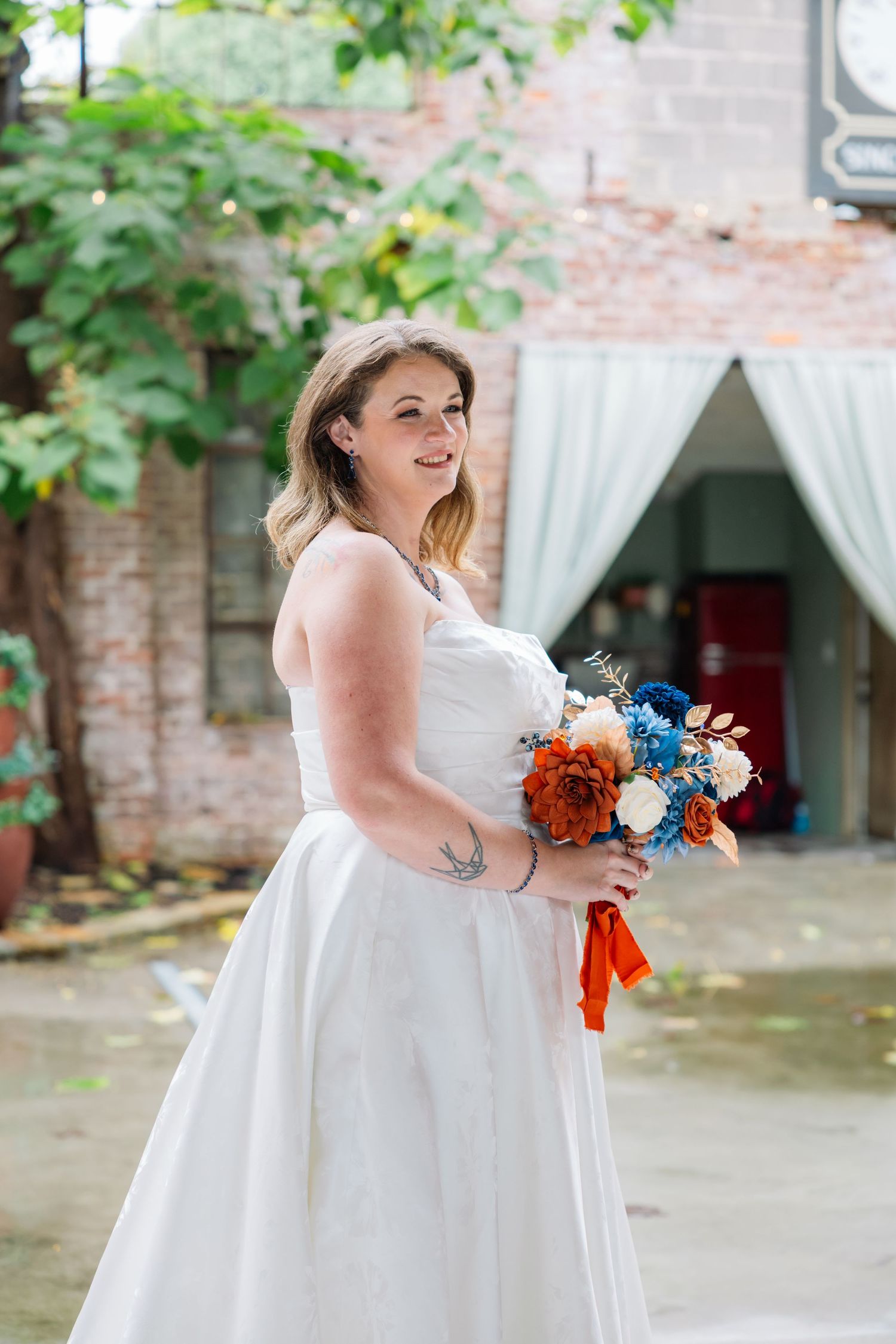 A bride in a white wedding dress holds a vibrant orange and blue bouquet while posing outdoors near a brick wall.