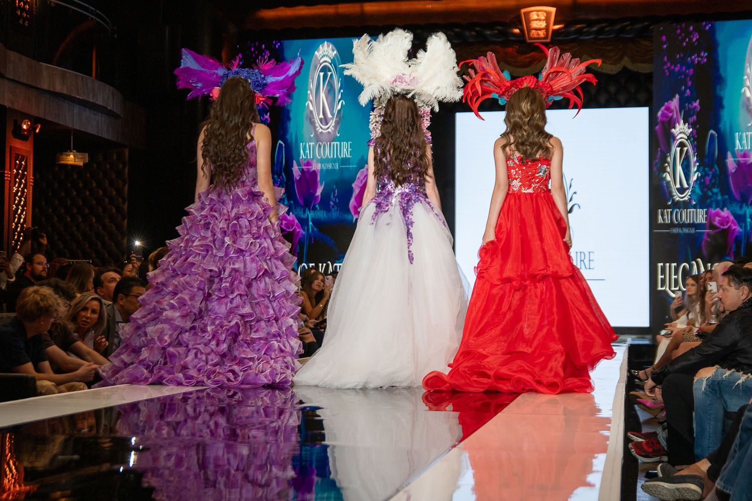 Three elegant ball gowns in purple, white and red displayed on a fashion runway with dramatic lighting and audience.