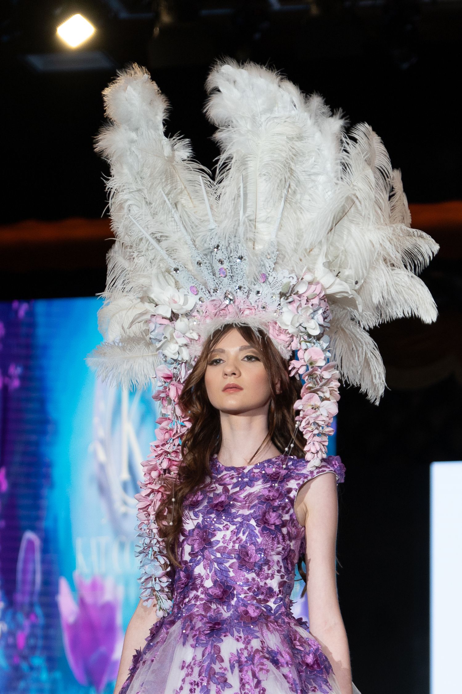 Model walks runway in dramatic purple gown with white feathered headpiece at fashion show.