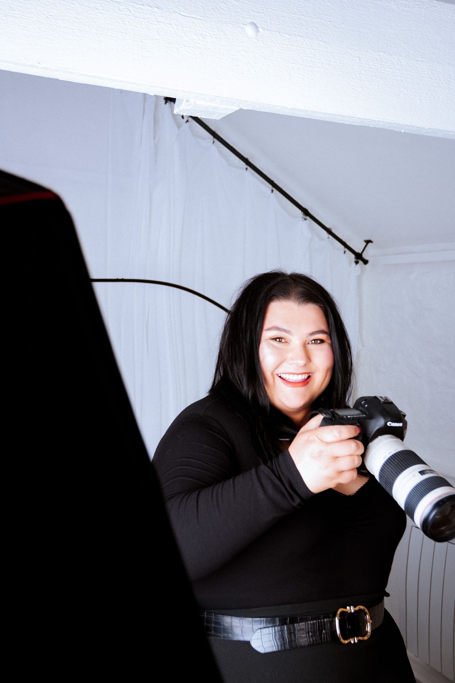 A photographer in black clothing smiles while holding a professional camera with telephoto lens.