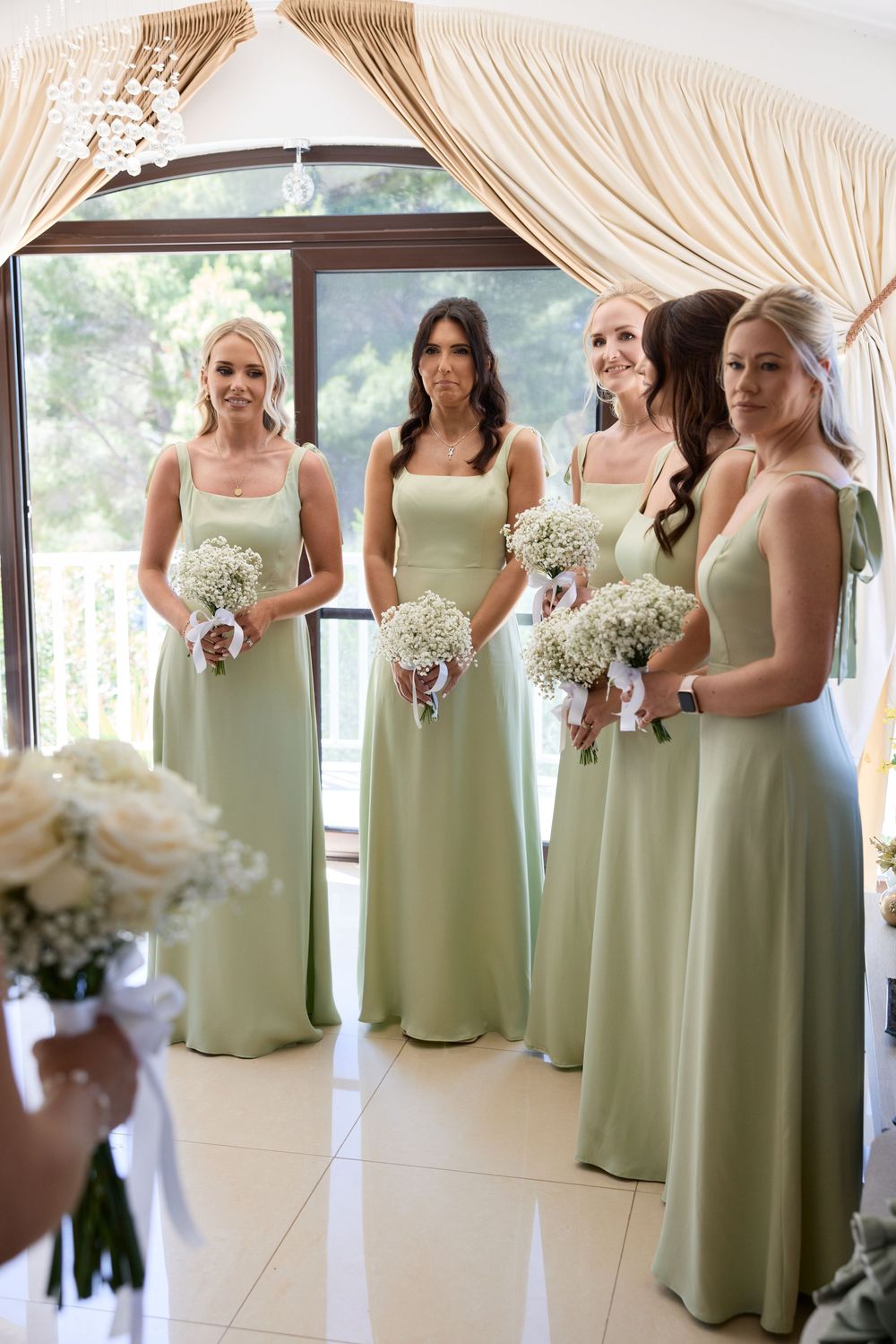 Bridesmaids in sage green dresses holding white bouquets in an elegant indoor setting.