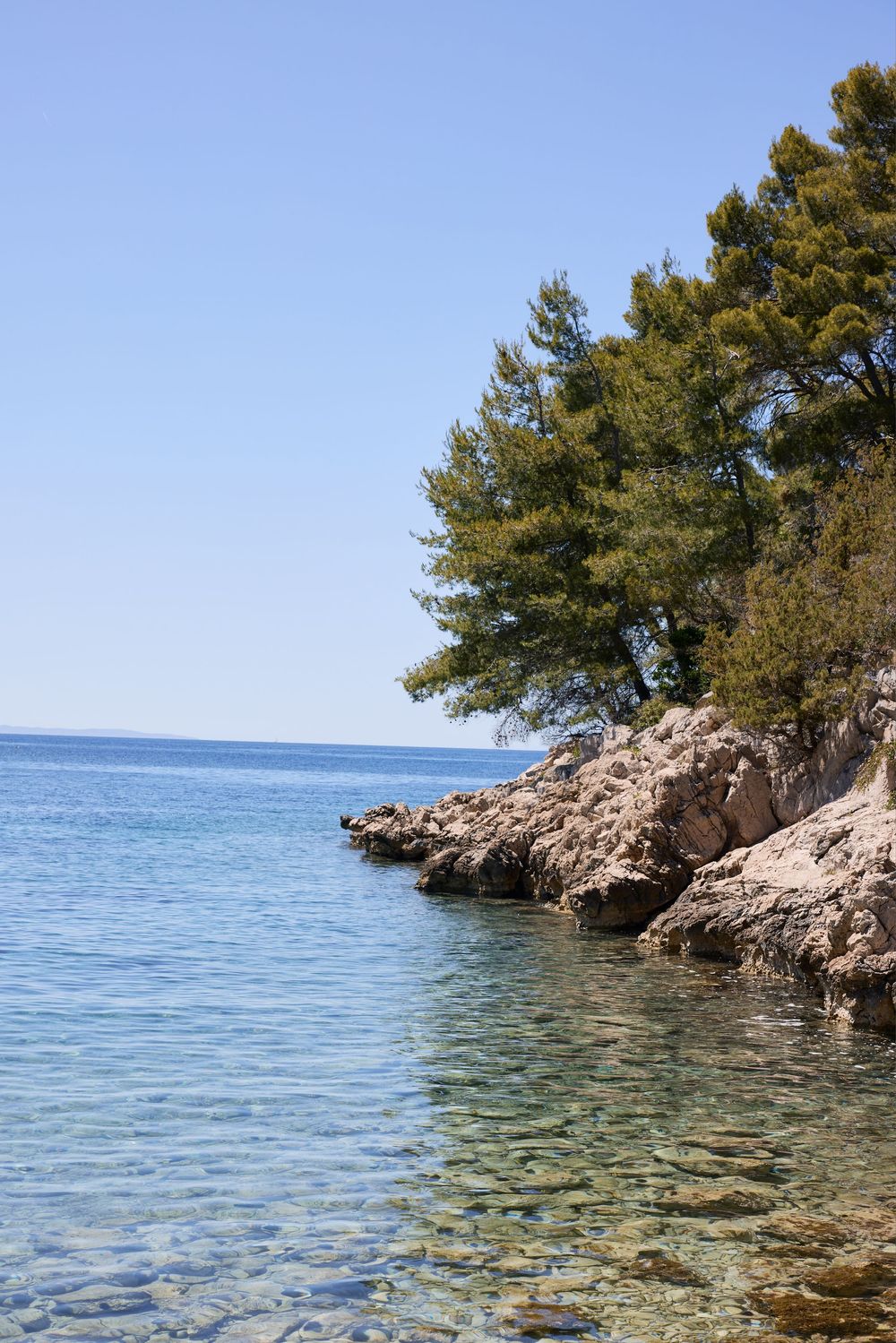 Crystal clear turquoise waters meet rocky coastline with pine trees in Mediterranean setting.