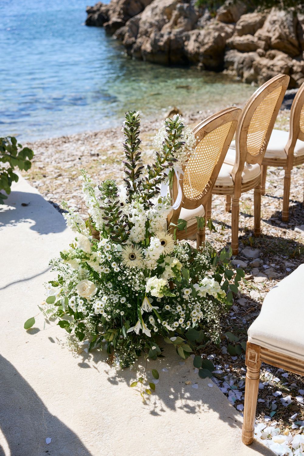White floral arrangements decorating wooden chairs on a rocky Mediterranean beach.
