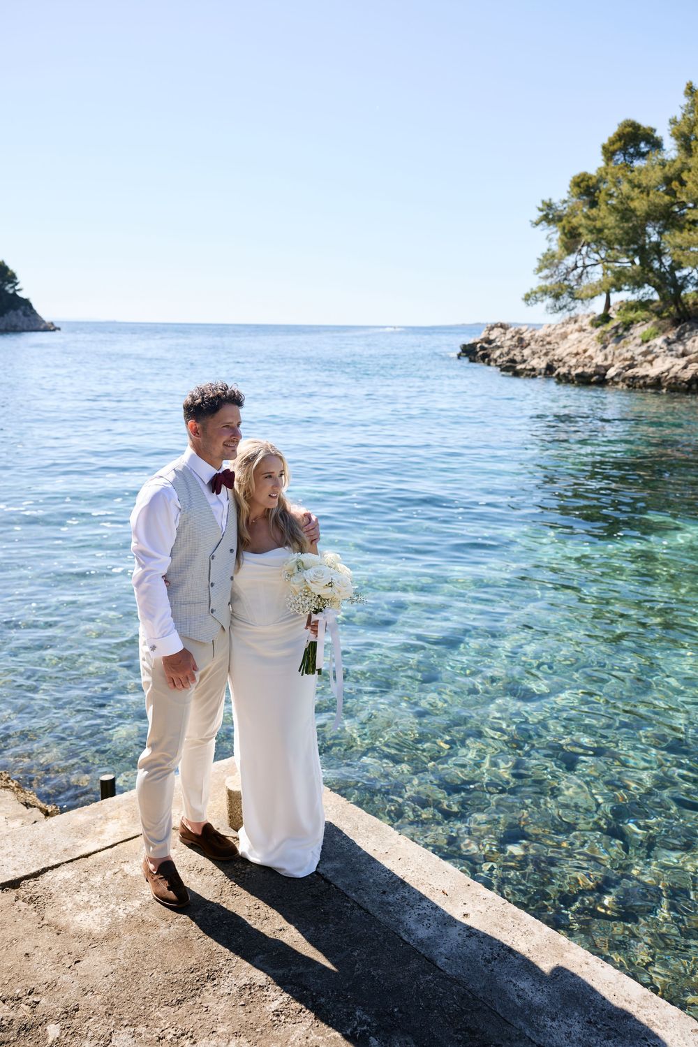 Couple in white wedding attire pose together on rocky Mediterranean coastline.