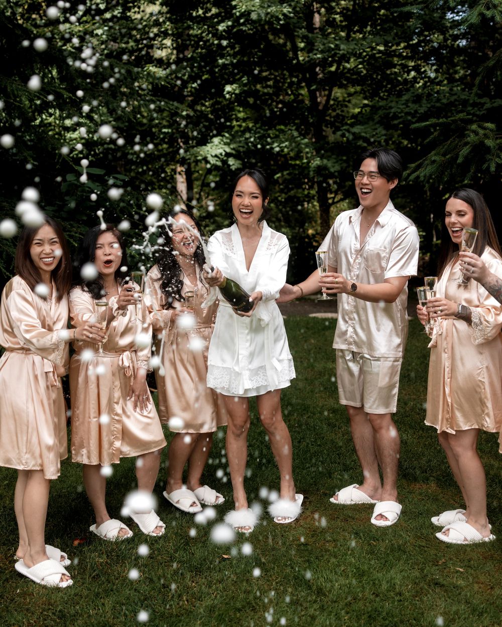 Wedding party members in matching silk robes pose playfully on grass for a group photo.