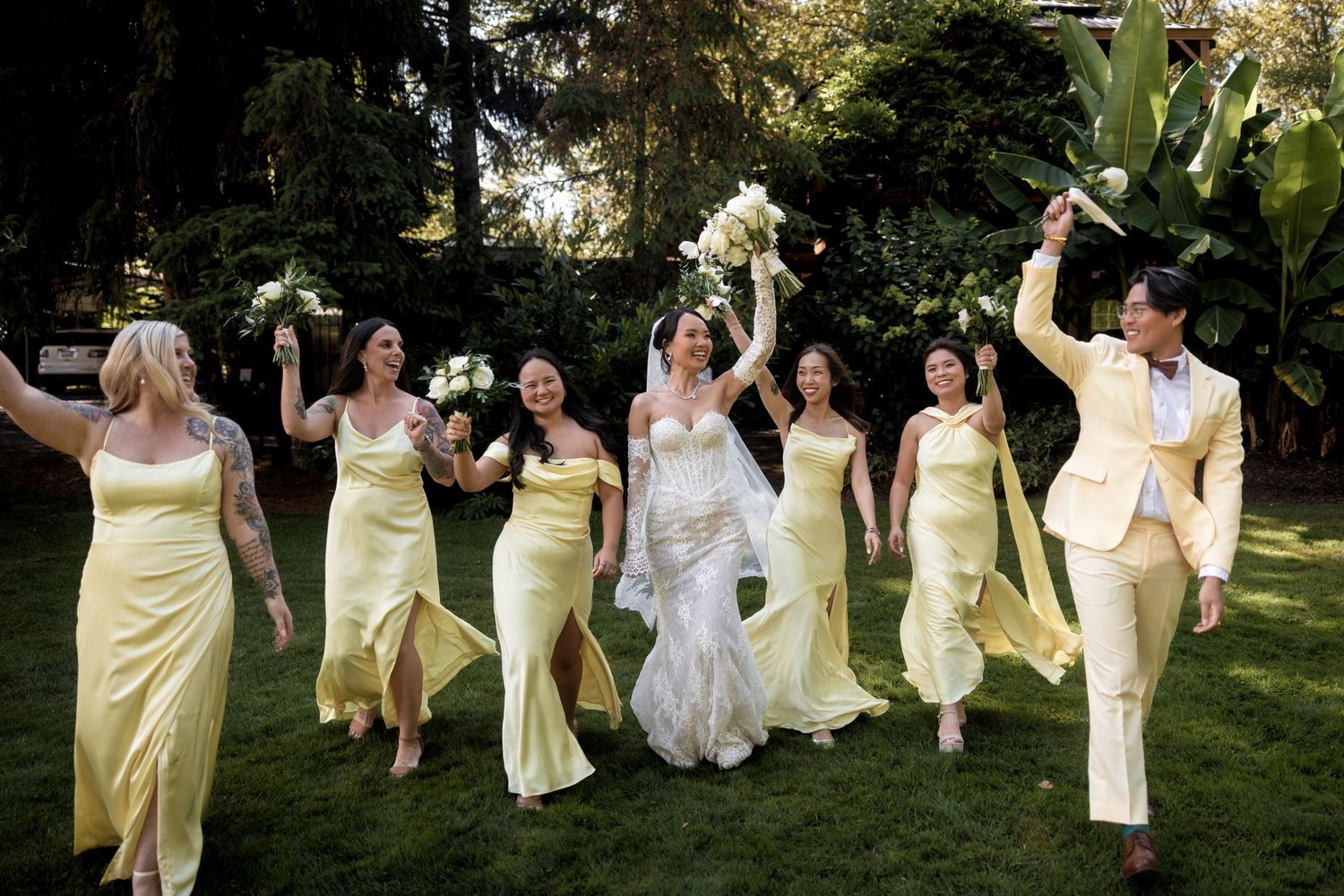 Wedding party in yellow and white dresses jumping joyfully in an outdoor garden setting.