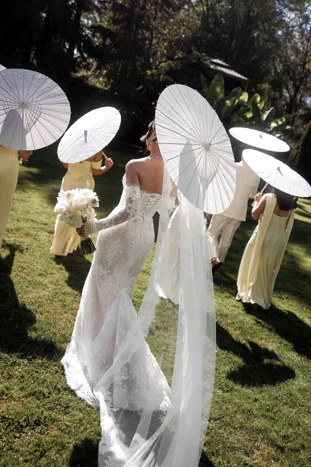 Wedding party poses with white paper parasols on a sunny lawn creating an elegant and ethereal scene.