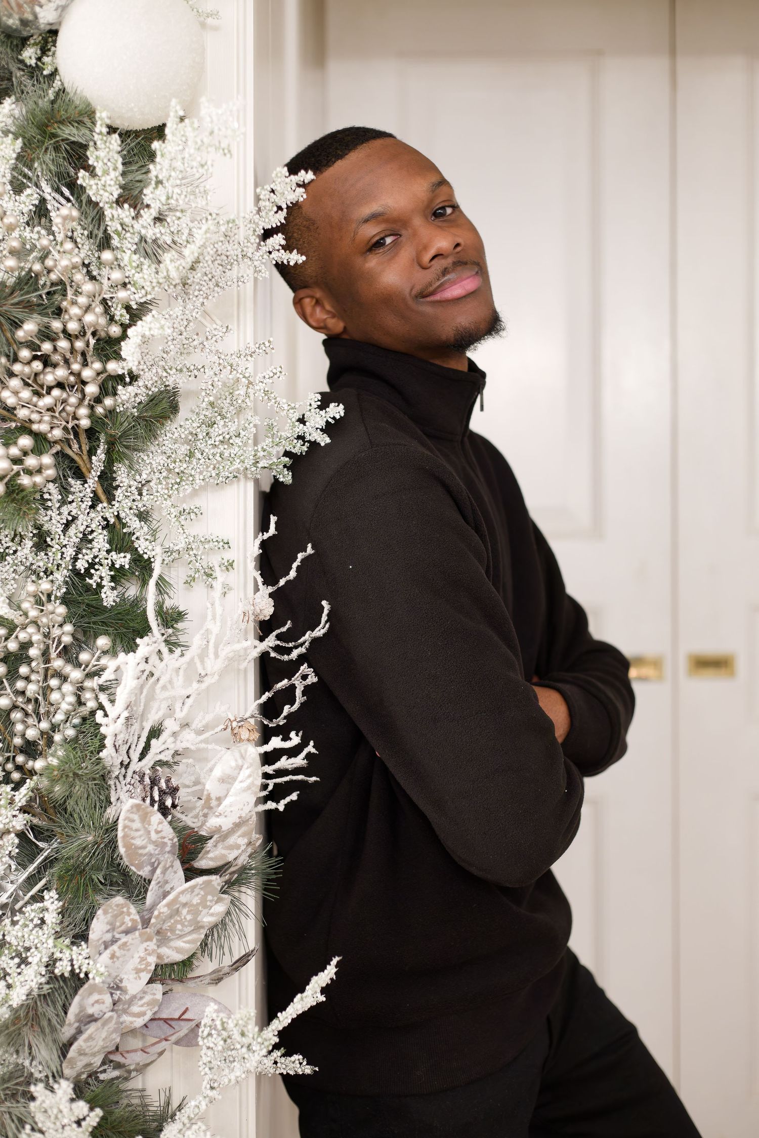 A young Black man in a black turtleneck leans against a wall beside elegant white Christmas decorations.