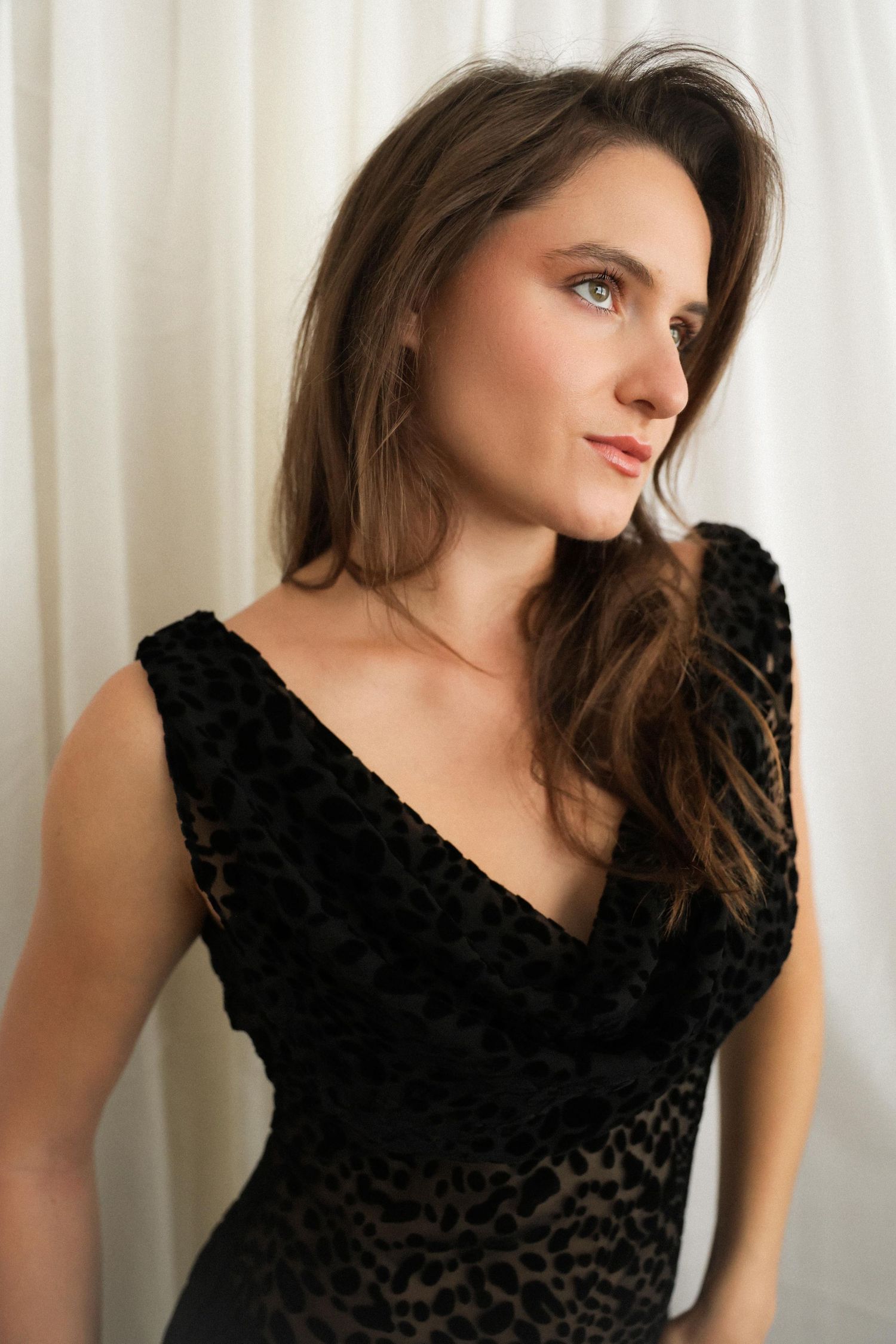 Portrait of a woman in an elegant black velvet dress posing against a light curtain background.