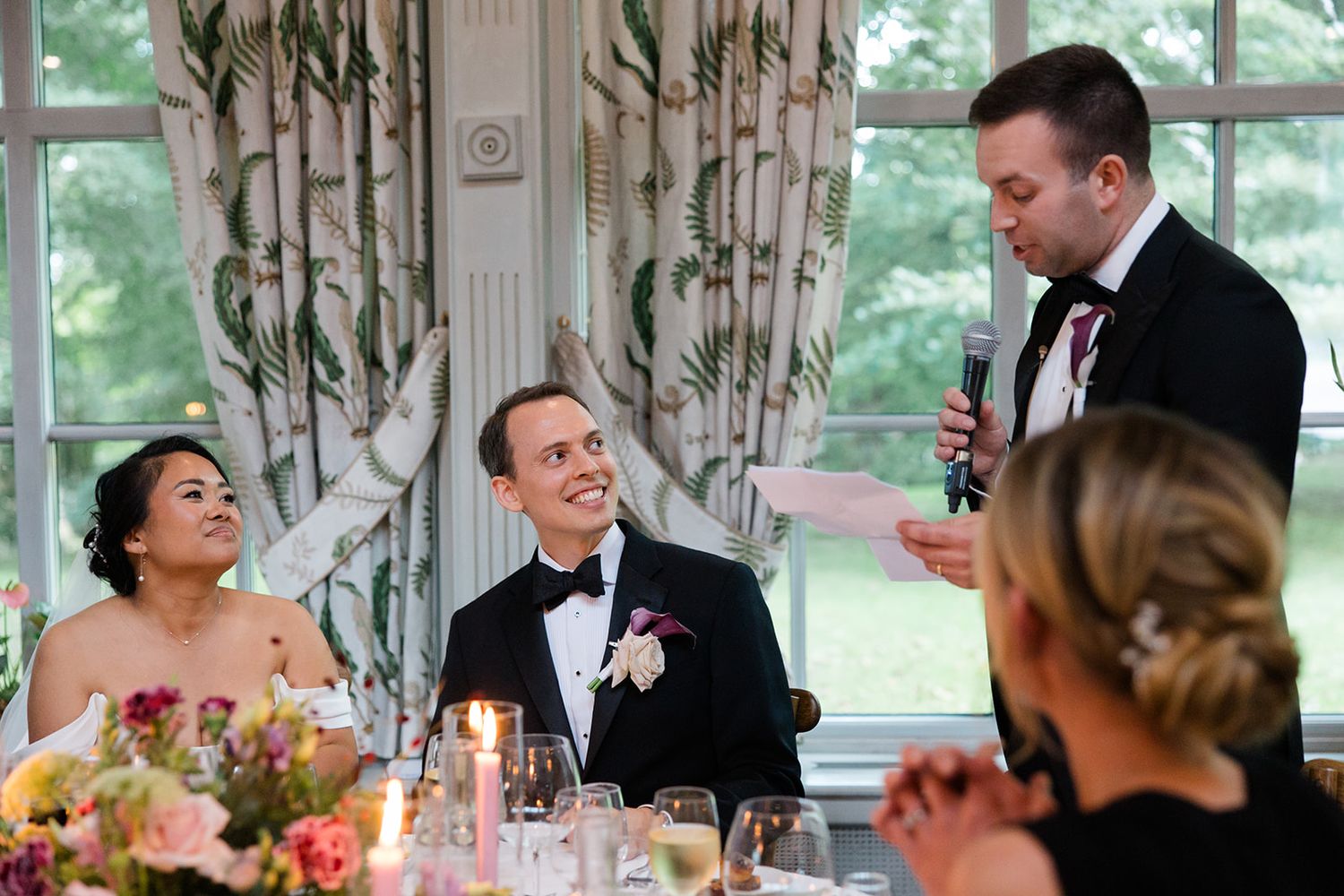 A candid moment at a wedding reception with guests seated at an elegant table with floral centerpieces and window views.
