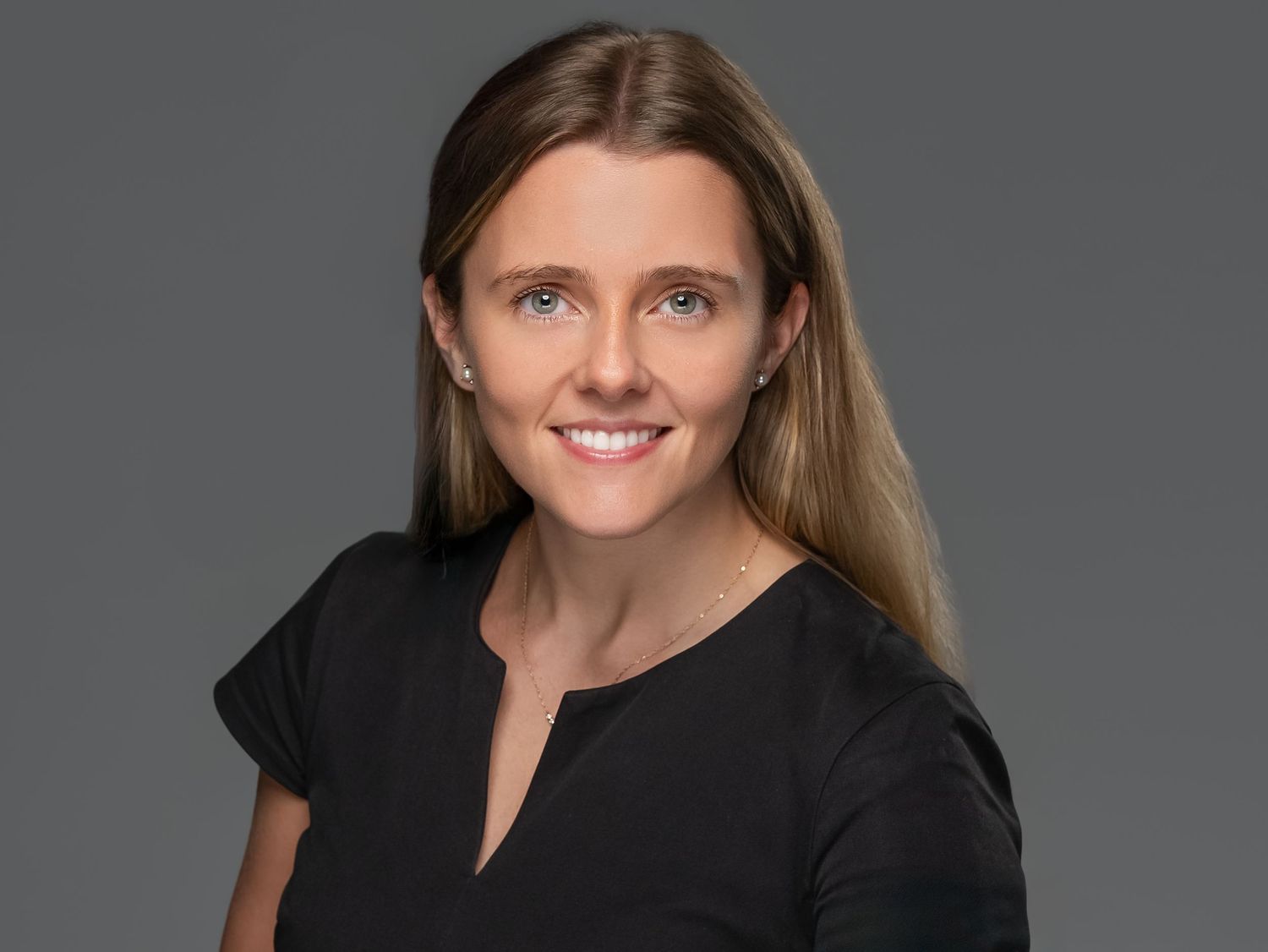 Professional headshot photo of someone with long blonde-brown hair wearing a dark colored top against a plain background.