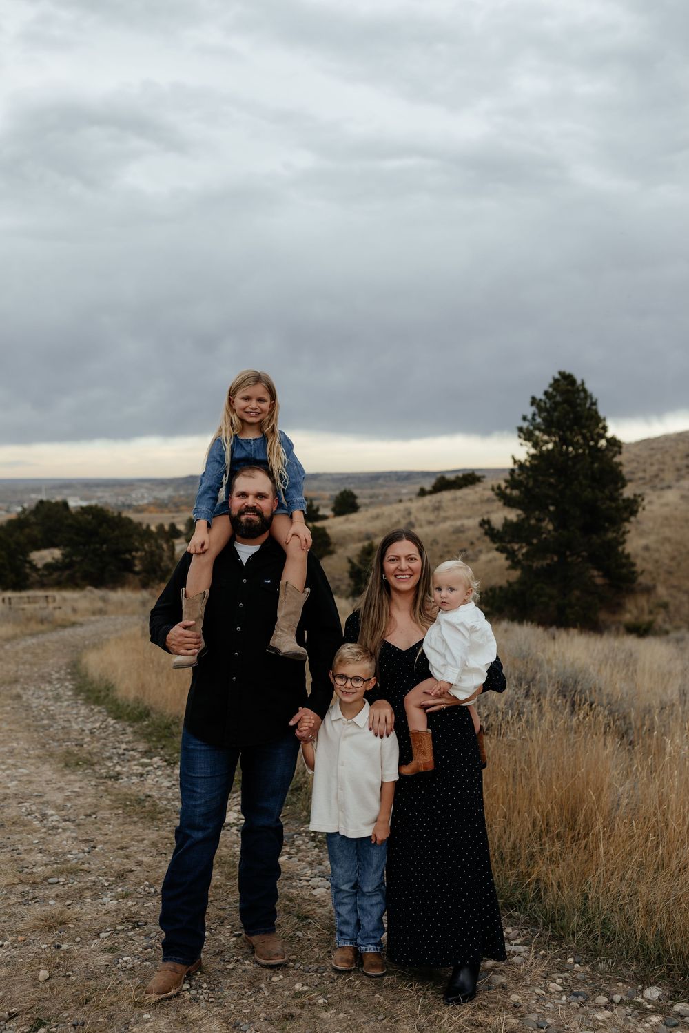 A family poses together on a rugged hillside path with dry grass and cloudy skies in the background.
