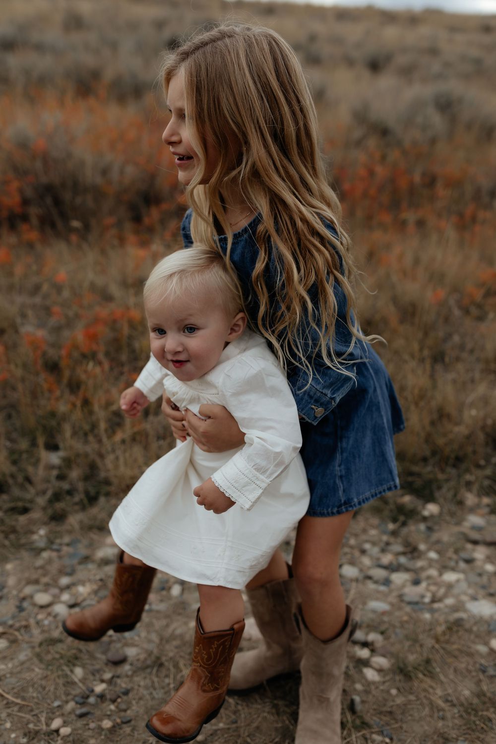 A young child in a white dress and brown boots stands outdoors with a caregiver in denim against an autumn landscape.
