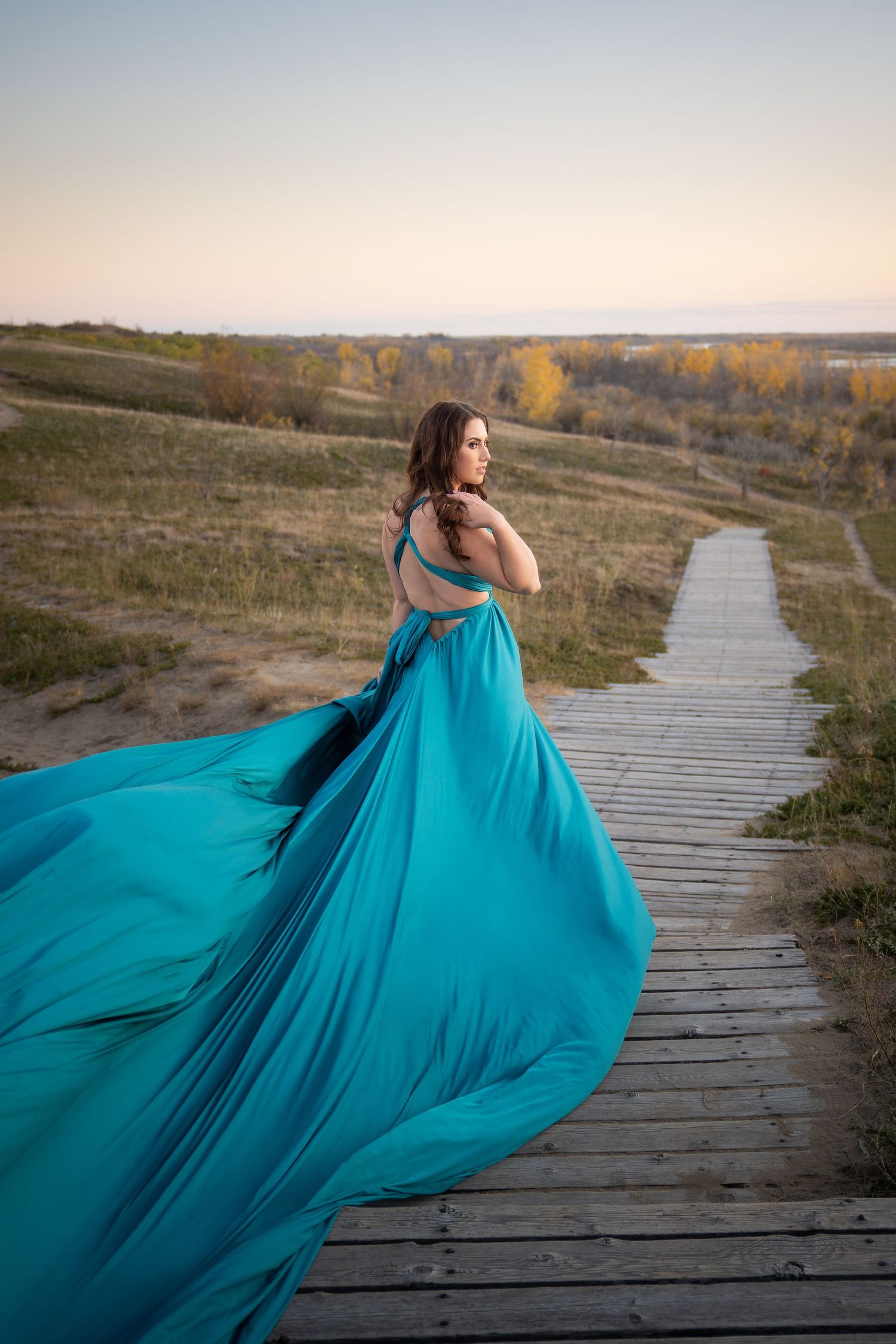 A flowing teal dress billows dramatically along a wooden boardwalk path at sunset in autumn.