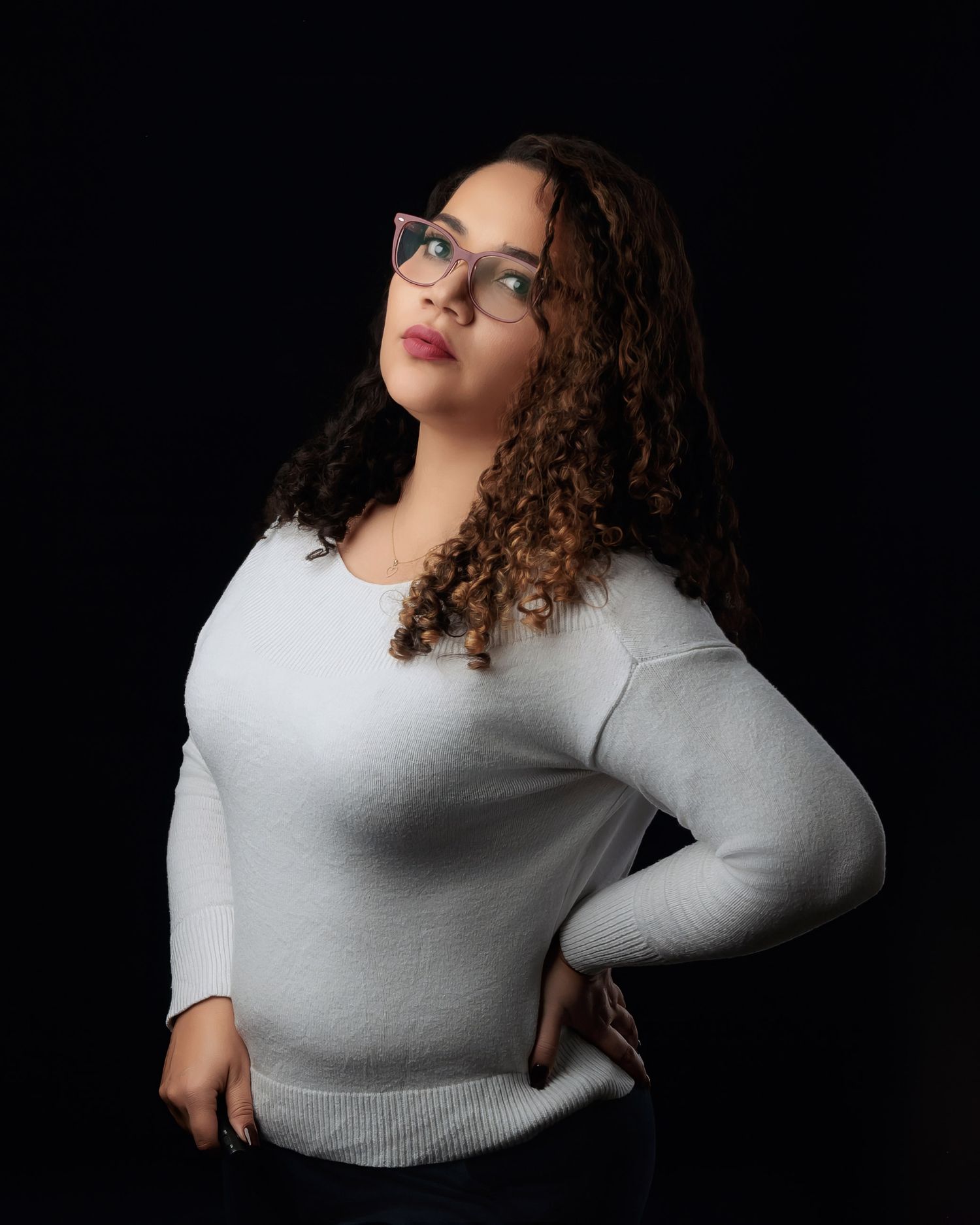 Young woman with curly hair and glasses poses confidently against a black studio background.