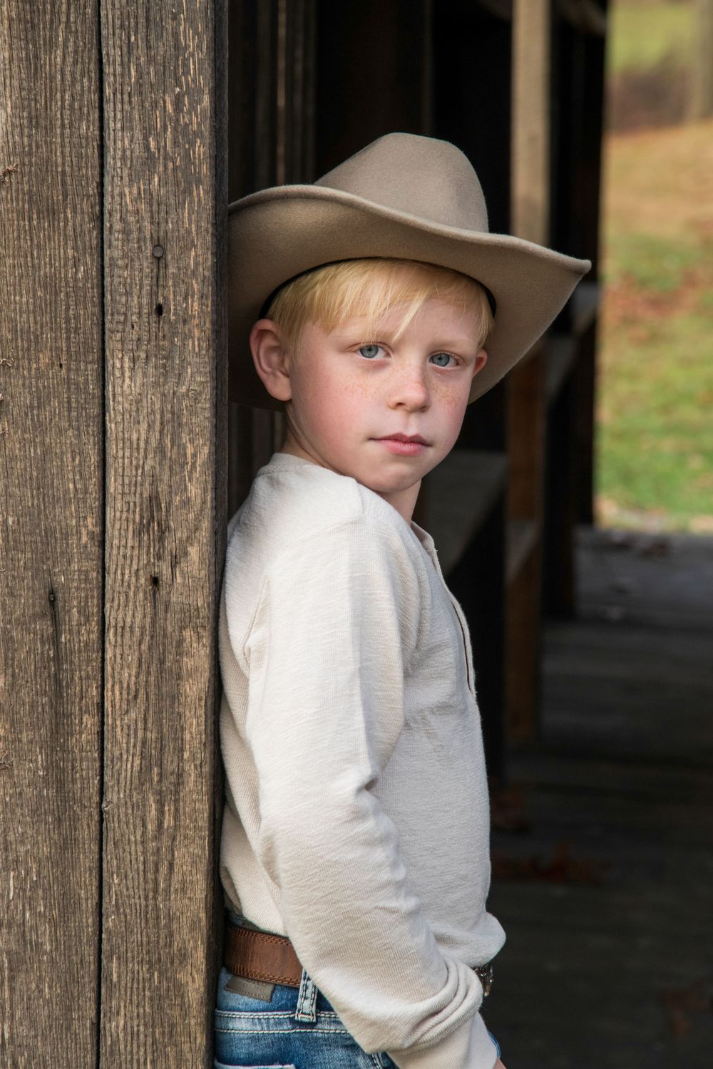 Serious young blond boy in cowboy hat and cream sweater leaning against a rustic wooden barn post.