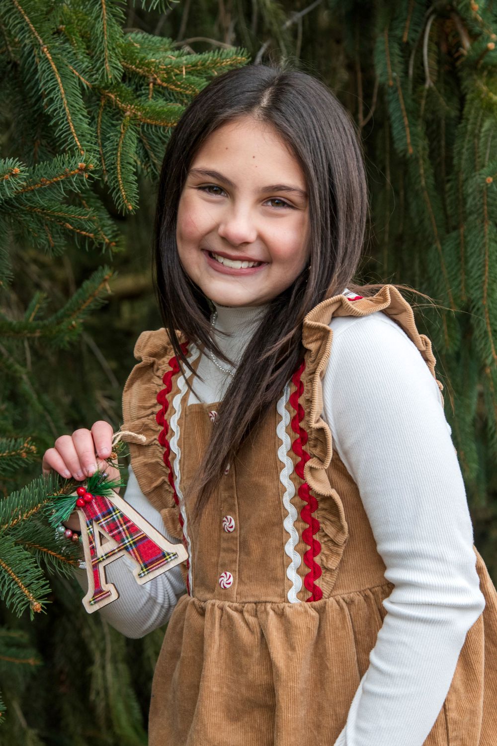 Happy young girl in brown ruffle dress holding a decorative letter A ornament beside a green Christmas tree.