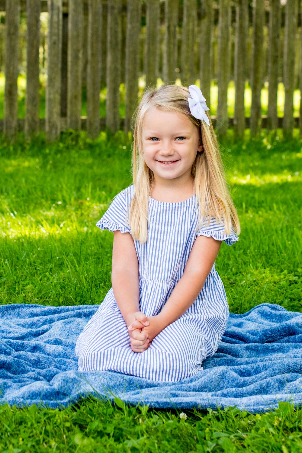 Cute blonde girl in striped dress smiles while sitting on a blue blanket in a sunny backyard.