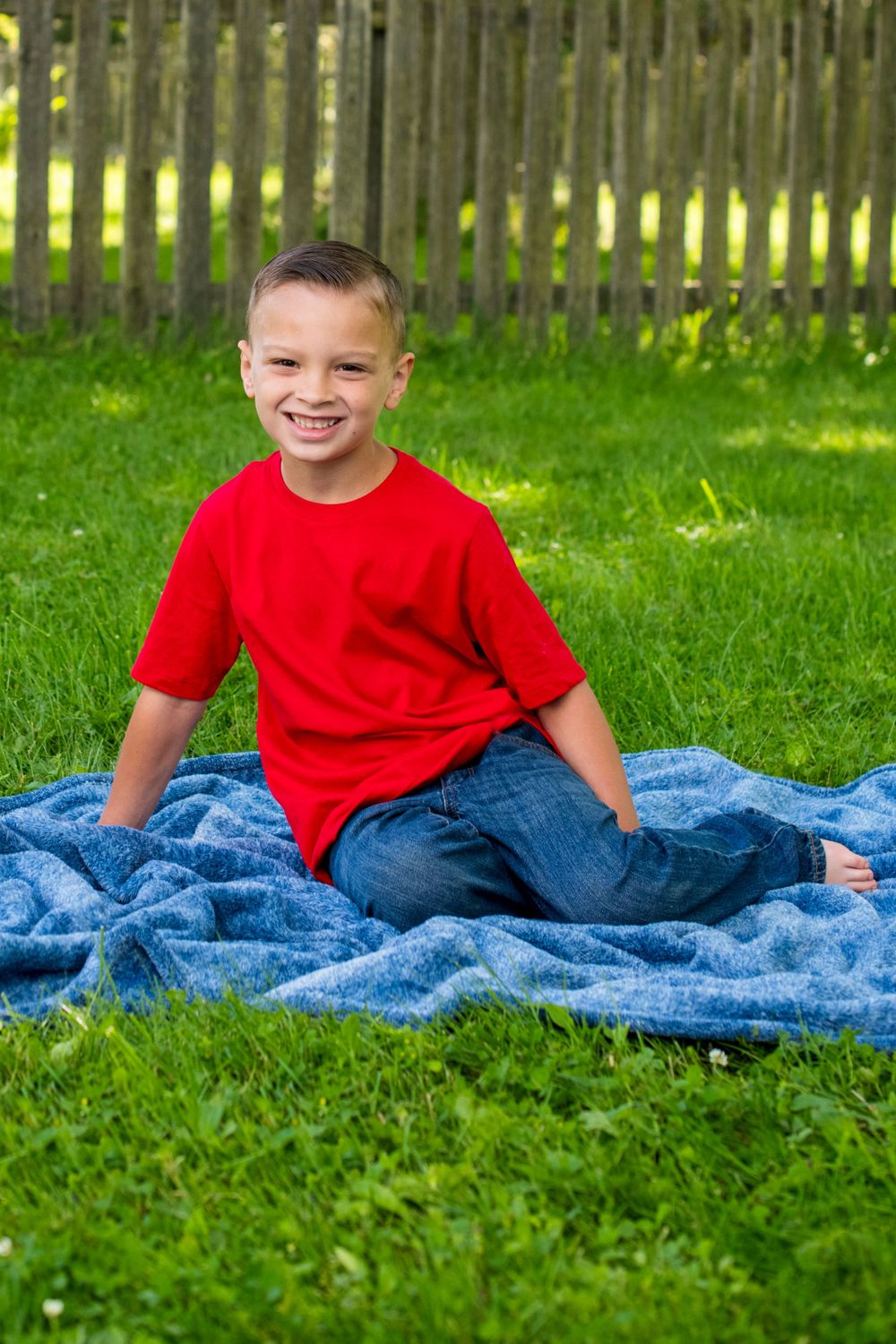 Happy young boy in red shirt sits on a blue blanket smiling in a bright green backyard.