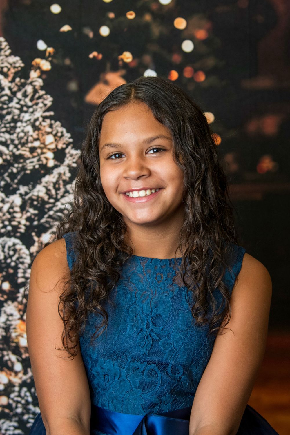 Smiling preteen girl with curly dark hair wearing a blue lace dress in front of bokeh Christmas tree lights.
