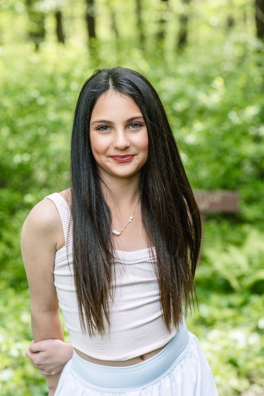 Young woman with long dark hair smiles outdoors, wearing a white crop top and skirt in a lush green forest setting.