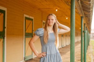 Girl in blue dress poses on rustic porch with wooden beams and green shutters in background.