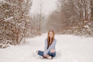 Winter senior portrait of person sitting cross-legged in snow-covered landscape.