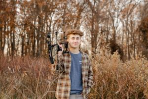 Photographer with professional camera poses in autumn field with tall grass and trees.