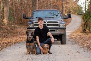 Person kneeling with two dogs in front of black pickup truck on forest road.