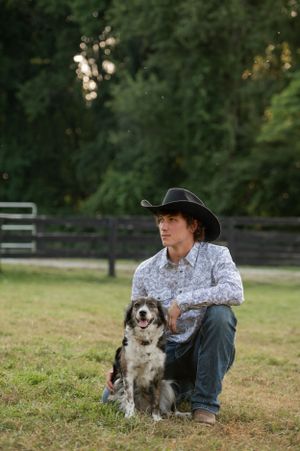 Person in cowboy hat and western shirt poses with friendly border collie dog on grassy field.