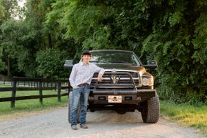 Person standing beside large lifted Dodge Ram truck on tree-lined country road.