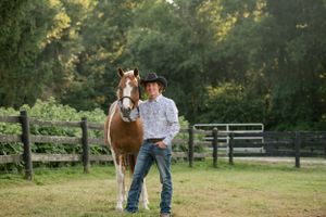 Person in western attire stands with beautiful brown horse in fenced pasture at sunset.