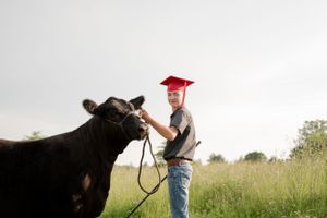 Graduate in red cap poses with black cow in grassy field.