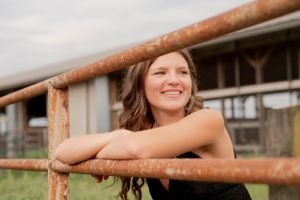 Person smiling while leaning on rustic metal farm fence during sunset.