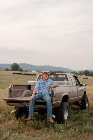 Person sitting on tailgate of lifted pickup truck in rural field setting at dusk.