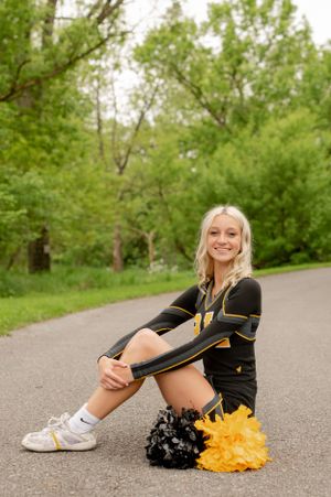 Cheerleader in black and gold uniform sitting with pom poms on paved path.
