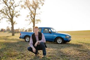 Senior photo of student sitting in front of blue pickup truck in rural field.