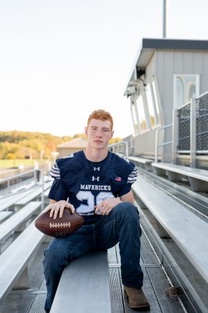 Football player in navy jersey sits on bleachers holding football at sunset.