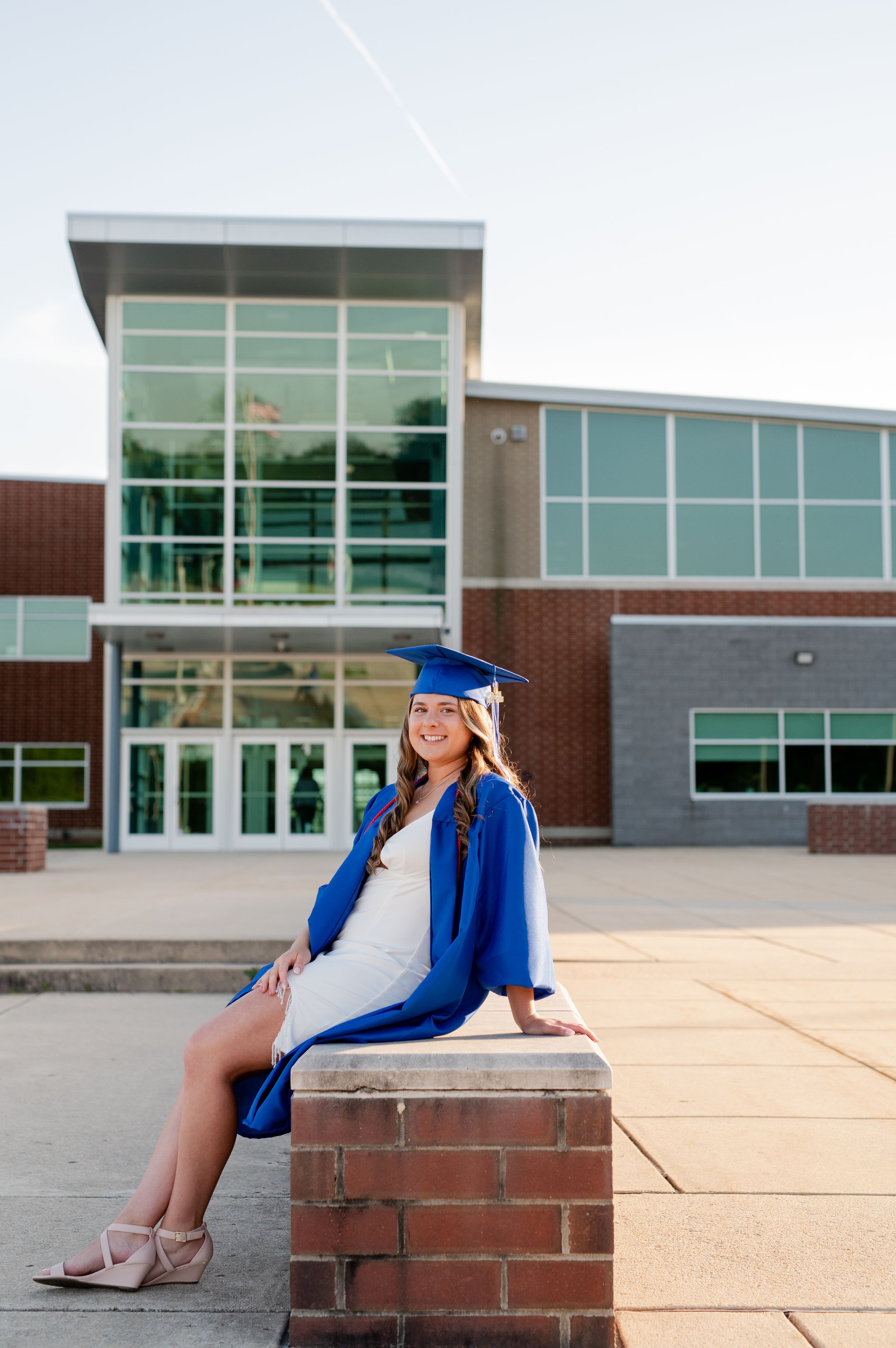 Senior in blue graduation gown sitting on brick wall outside modern school building.