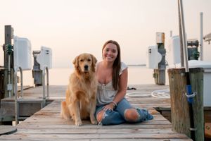 A golden retriever and person sitting together on a wooden dock at sunset near boats and water.