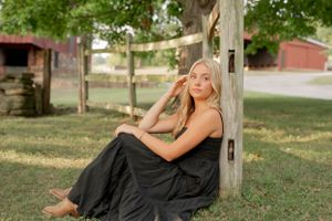 Person in a black dress posing gracefully against a wooden post in a sunny garden setting.