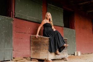 Person in a black dress seated on a rustic wooden box against a red barn wall.