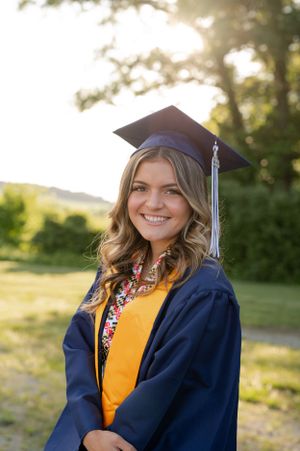 Graduate in navy cap and gown smiling outdoors on sunny day.