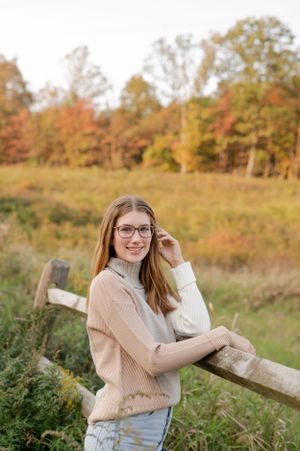 Person in cozy sweater leaning against wooden fence with autumn foliage in background.
