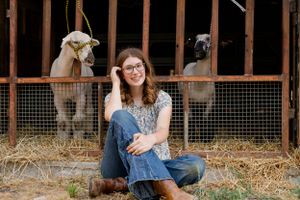 Person in denim sitting with goats in a barn stall.