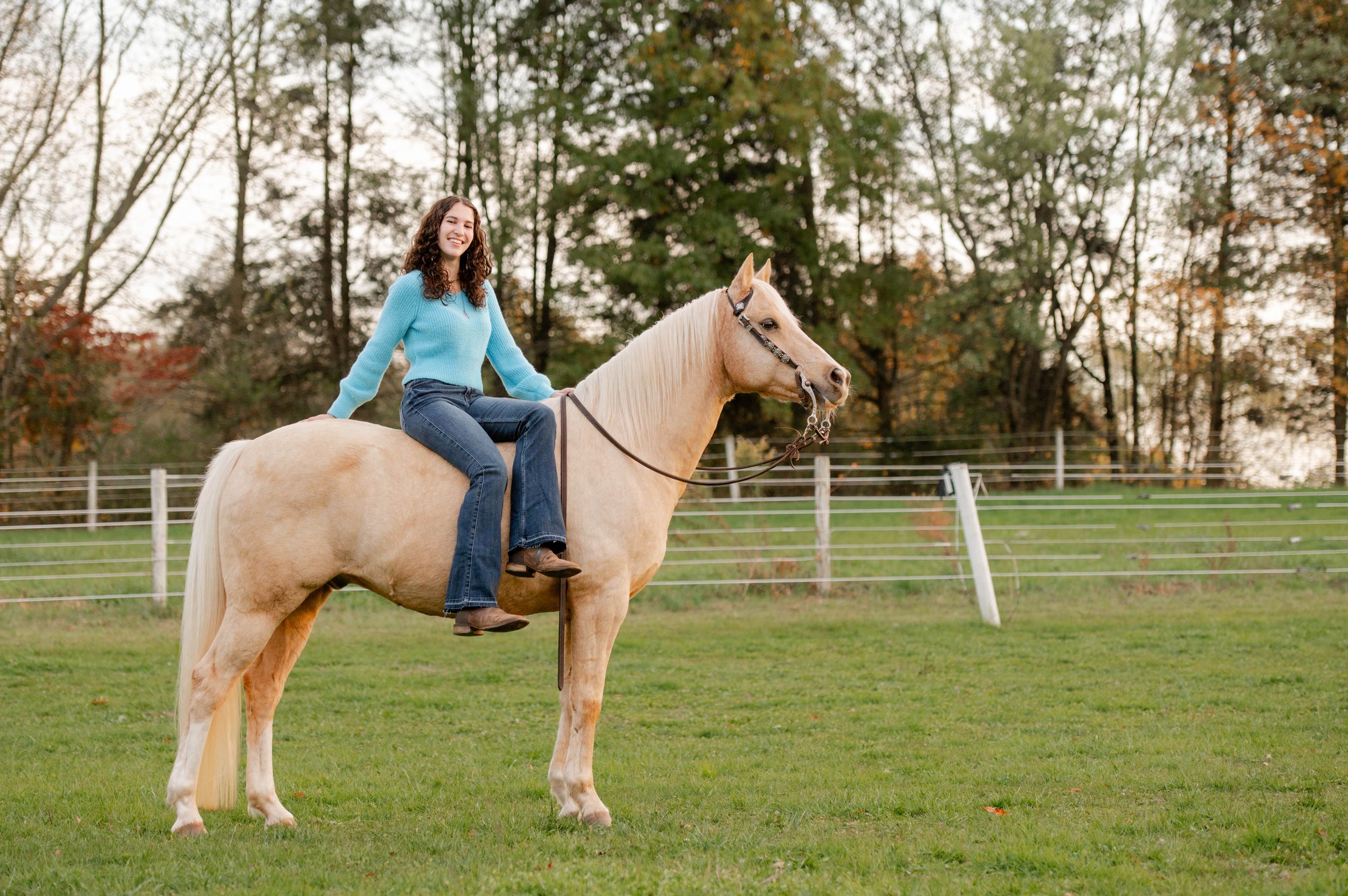 Rider sits atop a cream-colored horse in a fenced paddock during fall.